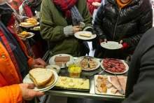 Photo : Des bénéficiaires tenant des assiettes avec du pain font la queue devant le chariot du petit-déjeuner, sur lequel il y a de la charcuterie et du fromage. Par respect pour la vie privée, aucun visage n'est visible.