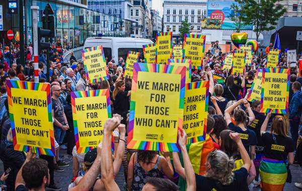Een groep mensen van achteren gefotografeerd terwijl ze meelopen in een mars van de Pride. Ze houden borden in de lucht met in een geel vlak in het zwart en in hoofdletters de woorden We march for those who can't. De borden zijn omlijnd in regenboogkleuren. 