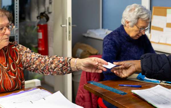 Photo : Une bénévole du service tickets vérifie une information dans sa farde tandis qu'une autre bénévole tend un ticket à un·e bénéficiaire. Par respect de la vie privée, le visage du ou de la bénéficiaire n'est pas visible.